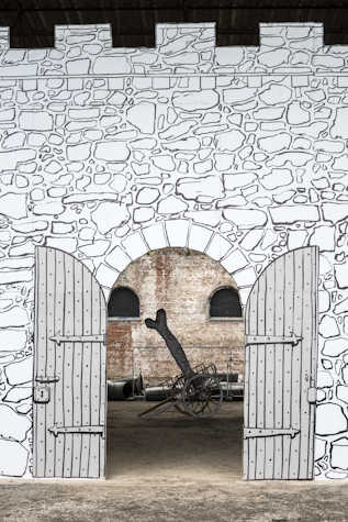 We can see a drawn black and white mural of a stone wall with an arched door in the middle, with wooden doors that are open to the sides. Through the door we can see a wheelbarrow that also looks drawn with a giant bone in it that has a message "to dog". Behind this is a real brick wall with two arched windows.