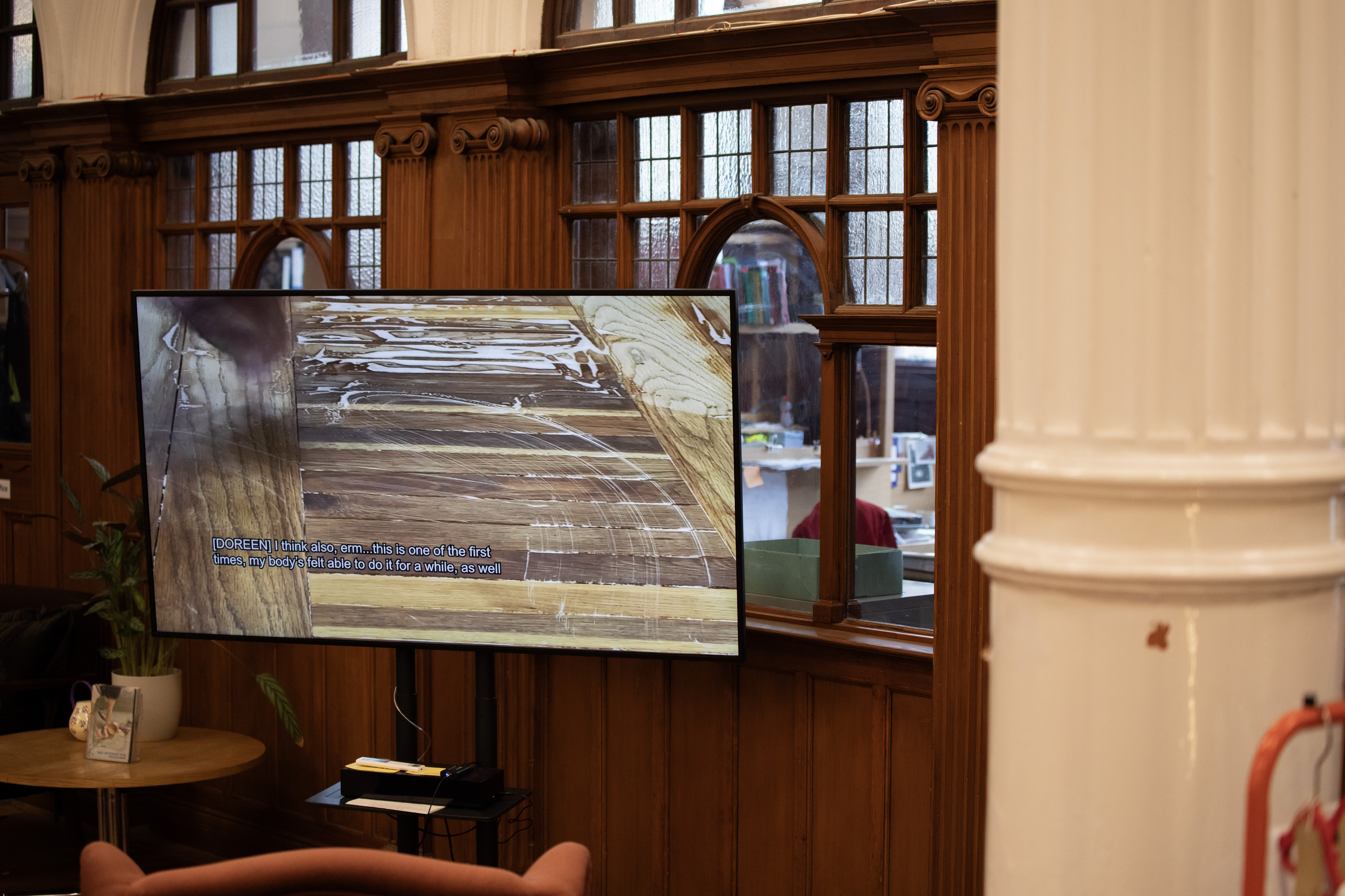 A screen in a wood-panelled room displays a close-up video of scarred wooden flooring, with subtitles reading "[DOREEN] I think also, erm...this is one of the first times, my body's felt able to do it for a while, as well.