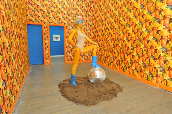 Front view of a figure wearing a swimming costume with one foot resting on a sphere. The walls of the interior have a patterned wallpaper. 