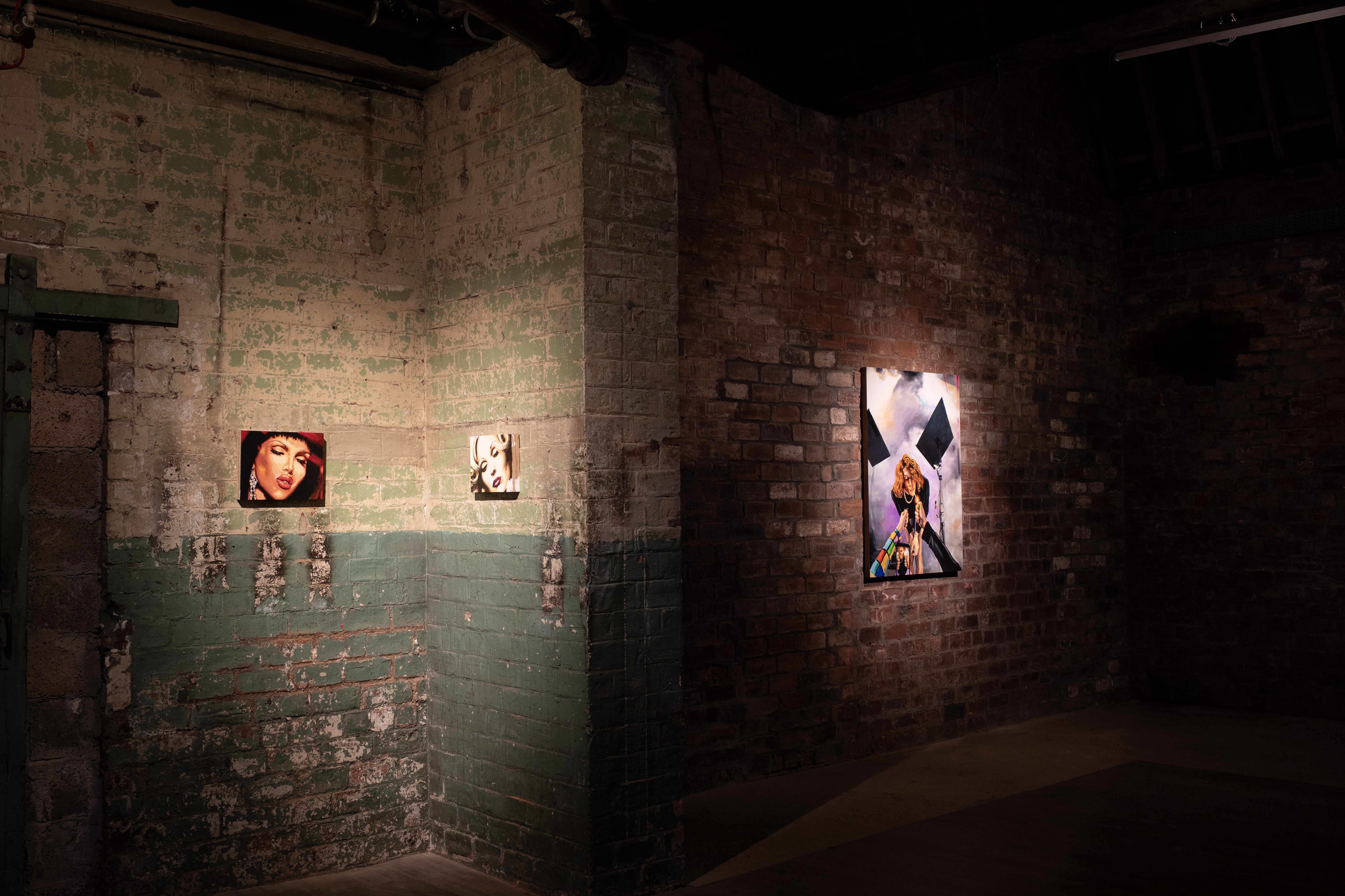 Gallery installation view of a painted corner, part-brick and part pale green-painted brick walls. Three small spotlit portrait paintings of women's faces are mounted at varying heights, with a larger canvas depicting a figure with black diamond-shaped props visible on the brick wall to the right.