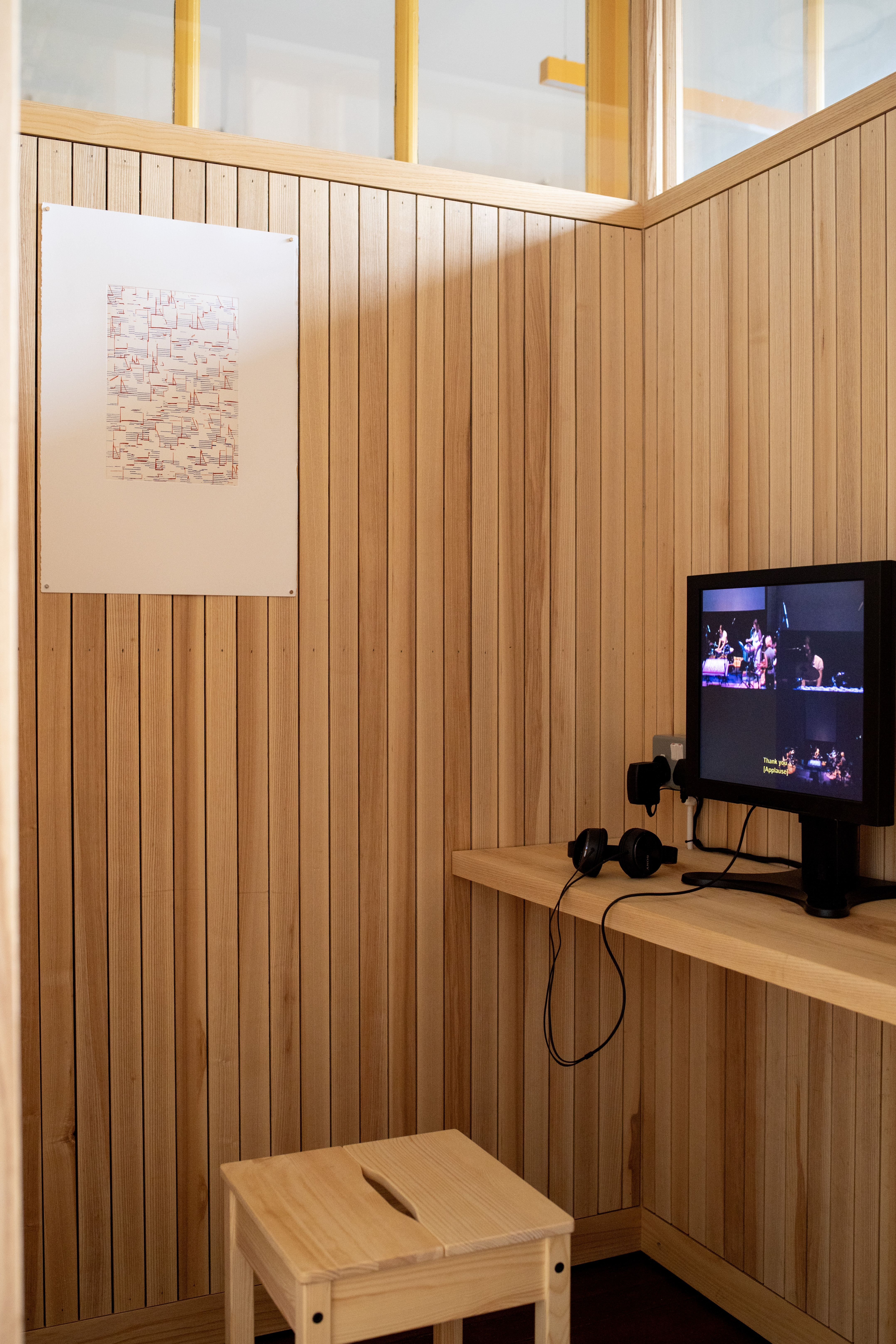 Corner of a wood-panelled booth with a large framed drawing of overlapping fine lines and marks on the left wall. A small CRT monitor on a shelf plays a video of musicians performing on stage, with headphones resting beside it and a wooden stool in the foreground.