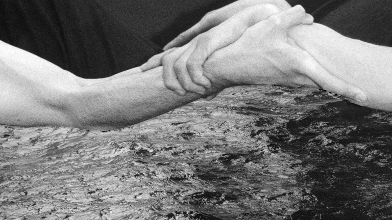 A black-and-white image shows two people holding on to one anothers arms. In the bottom of the photo is water or waves. 