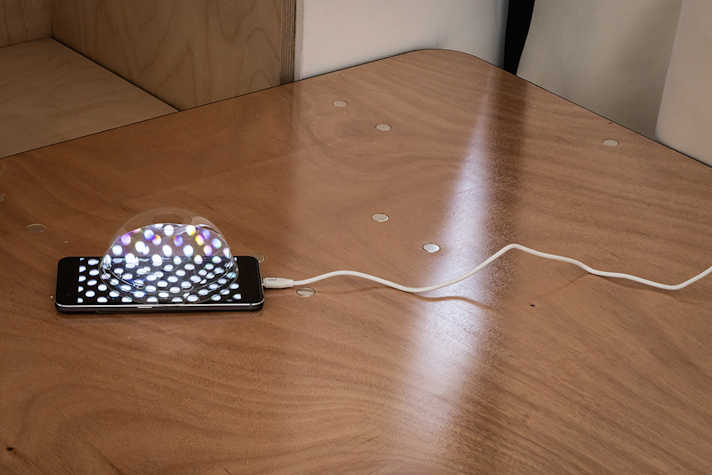 A phone displaying an image of white dots is displayed on a table with a cable connected to it. There is a spherical see-through shape on top of the phone screen.