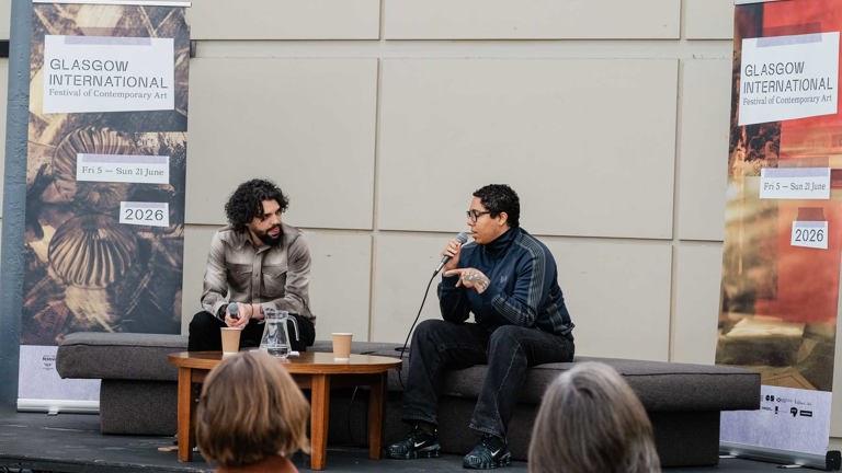 Two men in conversation on an outdoor stage, one gesturing while speaking into a microphone, the other listening, seated either side of a low table with water and coffee cups. Glasgow International banners flank the stage, and audience members are visible in the foreground.