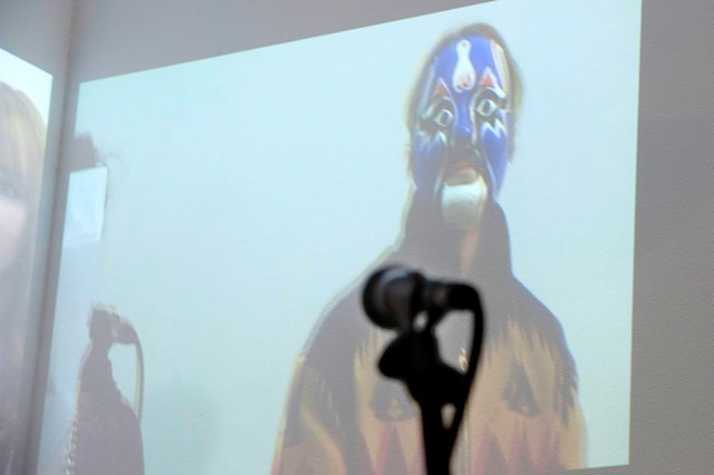 projection of a man wearing a wrestling mask. in front there is a microphone on a stand.
