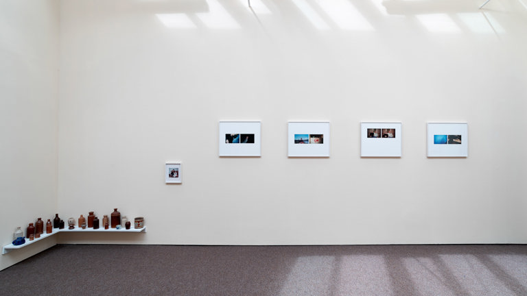 Installation view, photographic images on wall, small corner shelf just above floor, lined with ceramic bottles of various colours and sizes