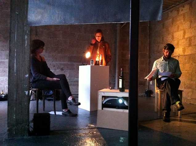 three people sat in a lowly lit room with brick walls. atop plinths there are lamps and glass bottles.