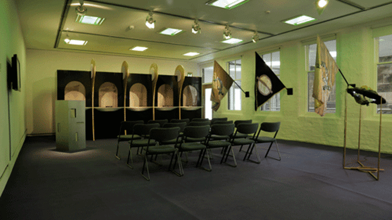 A room with some chairs arranged in rows facing a row of constructed alcoves. Flags on flag poles are fixed to the wall on the right. 