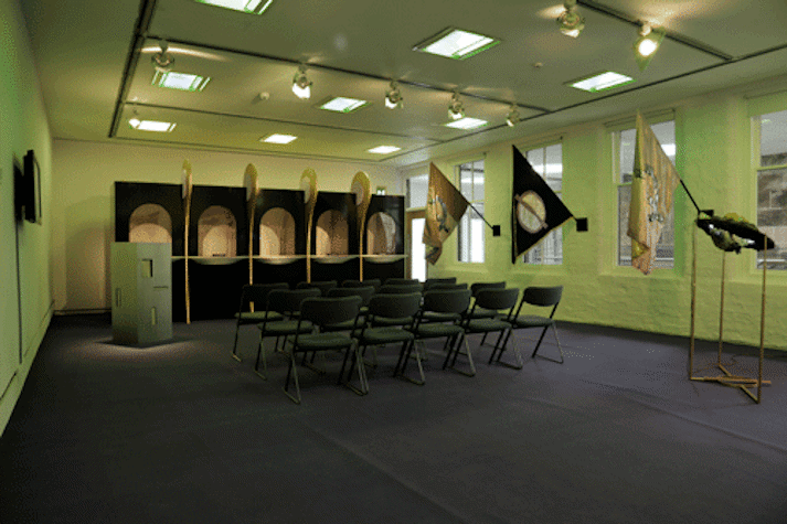 A room with some chairs arranged in rows facing a row of constructed alcoves. Flags on flag poles are fixed to the wall on the right.