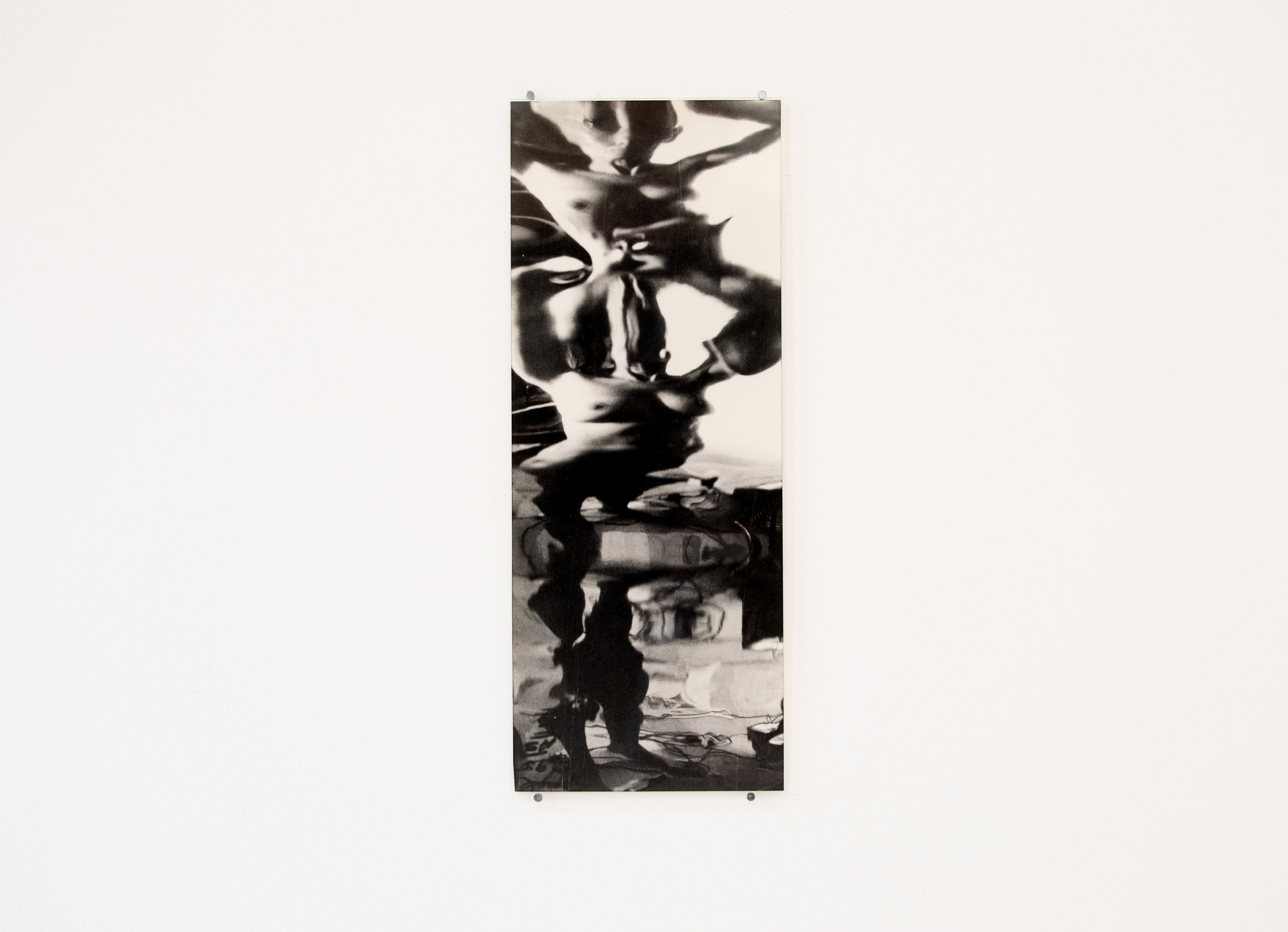 A single black-and-white photograph mounted on a white gallery wall, showing a warped, mirror-like reflection of a nude figure and interior objects.