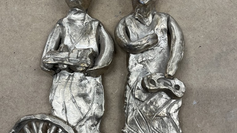 An image of two metal sculptural piece. Both depict people, the person on the left is standing behind a wheel, the one on the right stands behind an anvil and holds a tool.