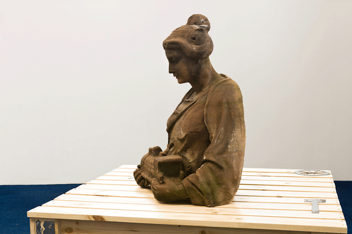 On a wooden structure is a section of a red sandstone sculpture showing the head and torso of a person with their hair drawn into a bun on their head and cradling a small train in their arms