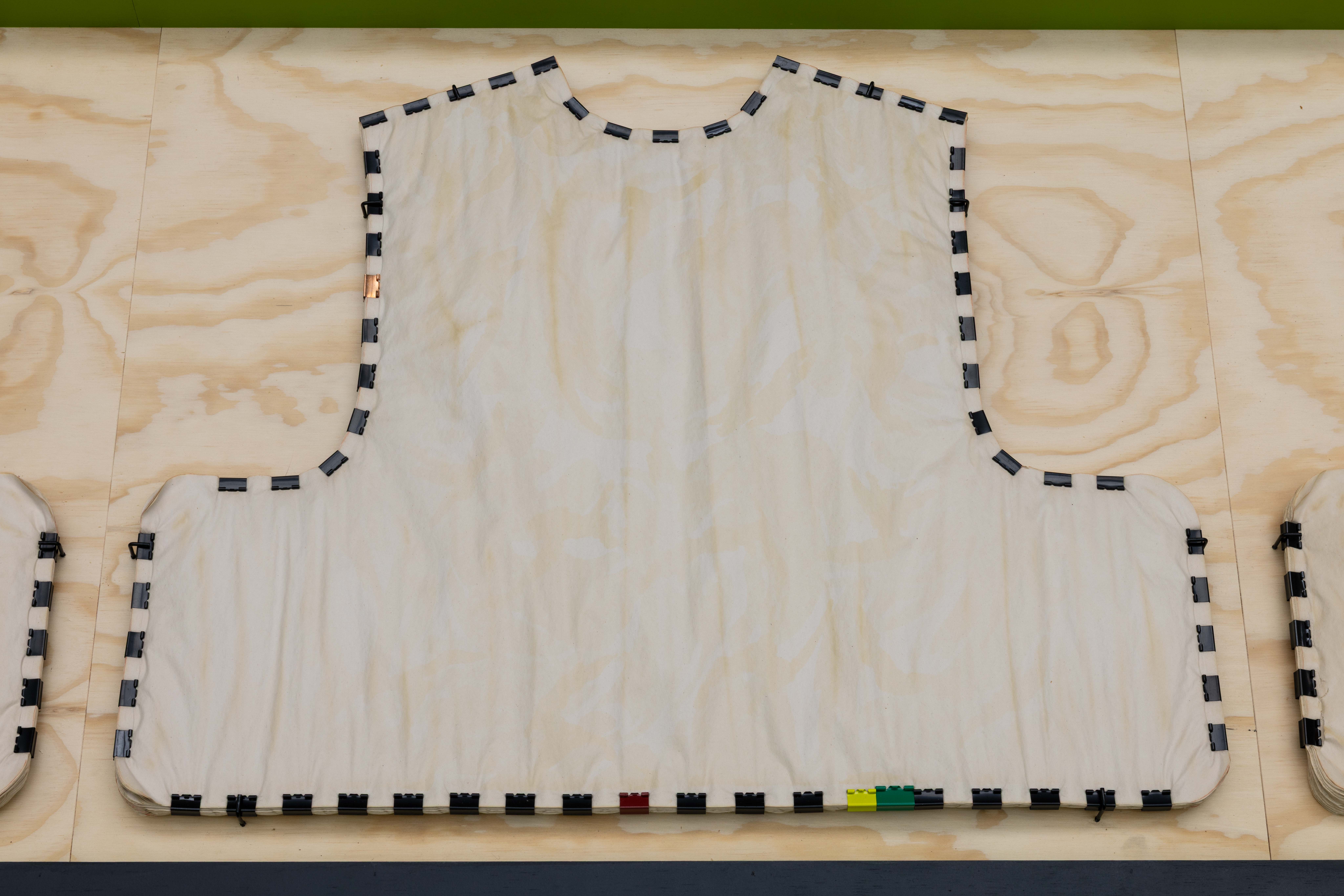 Close-up detail of a vest-shaped fabric form mounted on plywood, its edges lined with small black plastic clips and a few coloured toy brick-like fasteners in red, yellow and green.