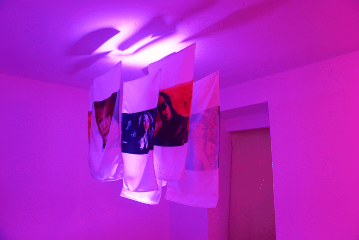 A room with pink neon lights has four fabrics hanging from the ceiling with images of women on it as a part of E. Jane's installation in Kelvin Hall as part of Glasgow International 2018.