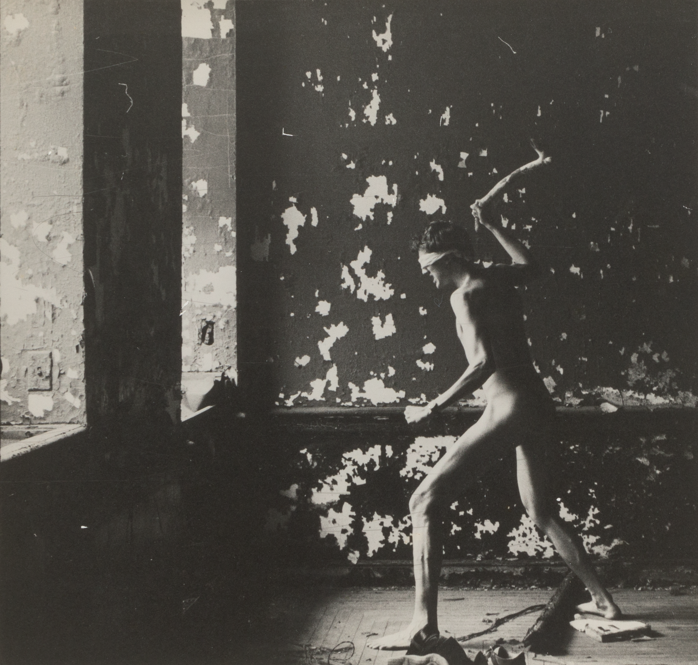 A black and white photograph of a blindfolded naked  person facing two large windows holding a stick above their head  like a weapon. They are in a room in an abandoned building with  debris on the floor and paint peeling from the walls.