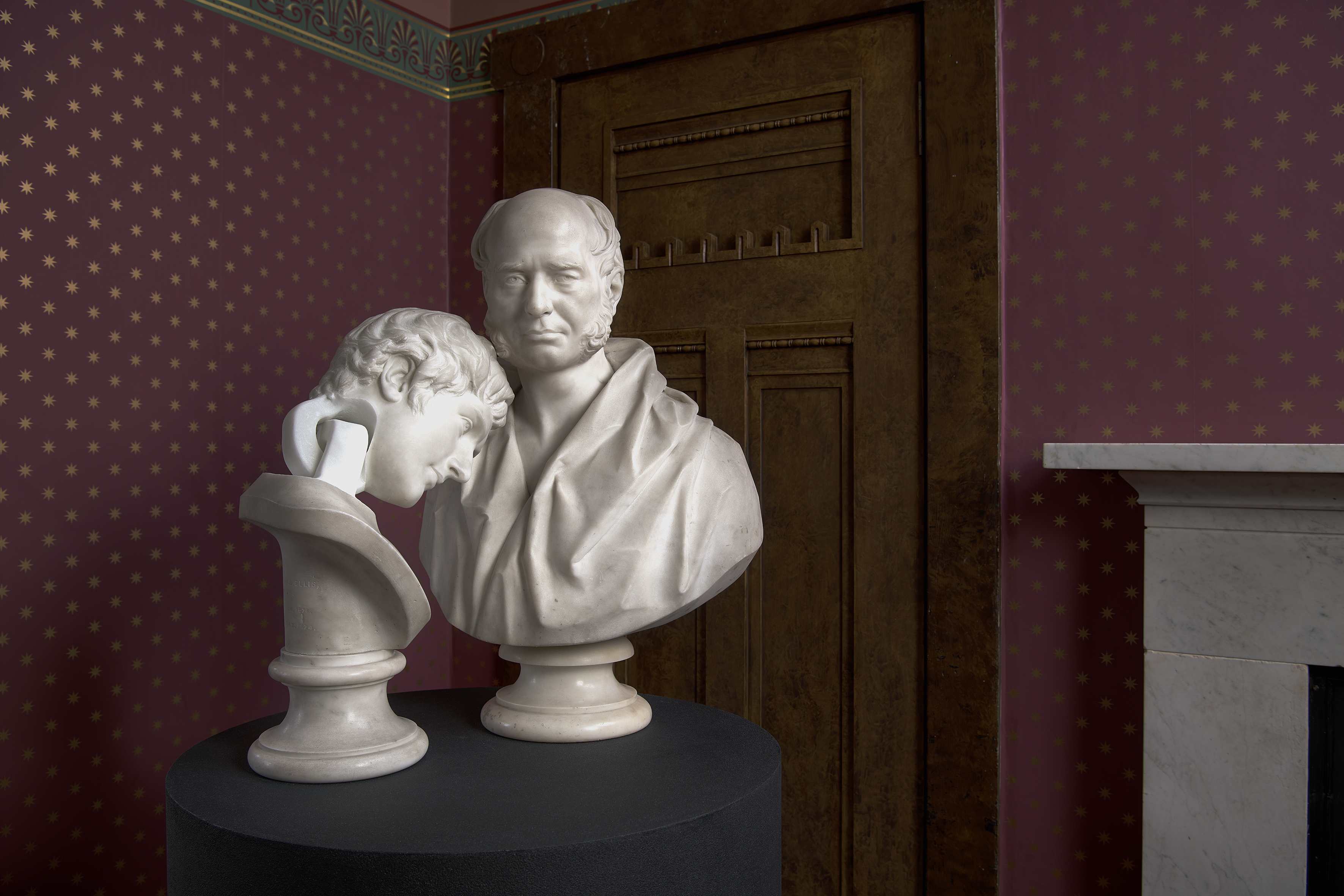 A row of classical marble busts, including a double bust of two leaning heads in the foreground, displayed in a bay window of Holmwood House's ornately papered room, part of Jon Owen's installation.