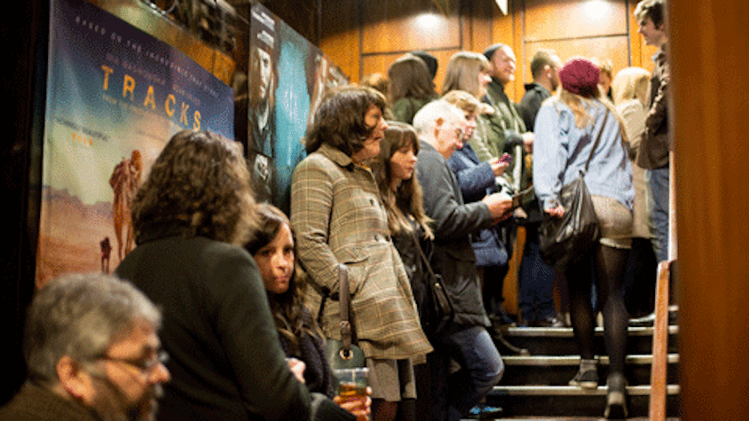 A queue of people are standing on the let side of a staircase, which is leading up to another floor. It has wooden railings on one side and posters of films on the left side, A woman in a hat is going up the stairs on the empty side of the staircase,