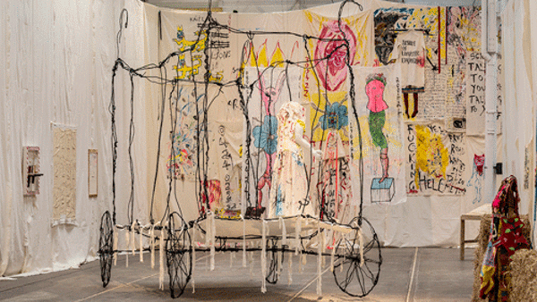 A wheeled carriage made from thin wire, displayed in a room filled with artwork displayed on fabric wall hanging.