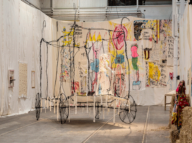 A wheeled carriage made from thin wire, displayed in a room filled with artwork displayed on fabric wall hanging.