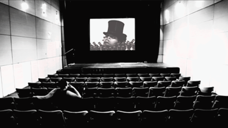 screen shot: image of the inside of a screening room, with an individual sitting on one of the chairs viewing the screen in front showing an individual wearing a hat 