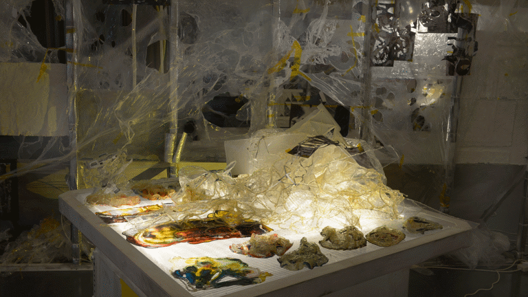 A table with light coming from within and different materials of different textures, colours and viscosity in web-like structures as part of an art installation in the Gallery of Modern Art, Glasgow International 2018.