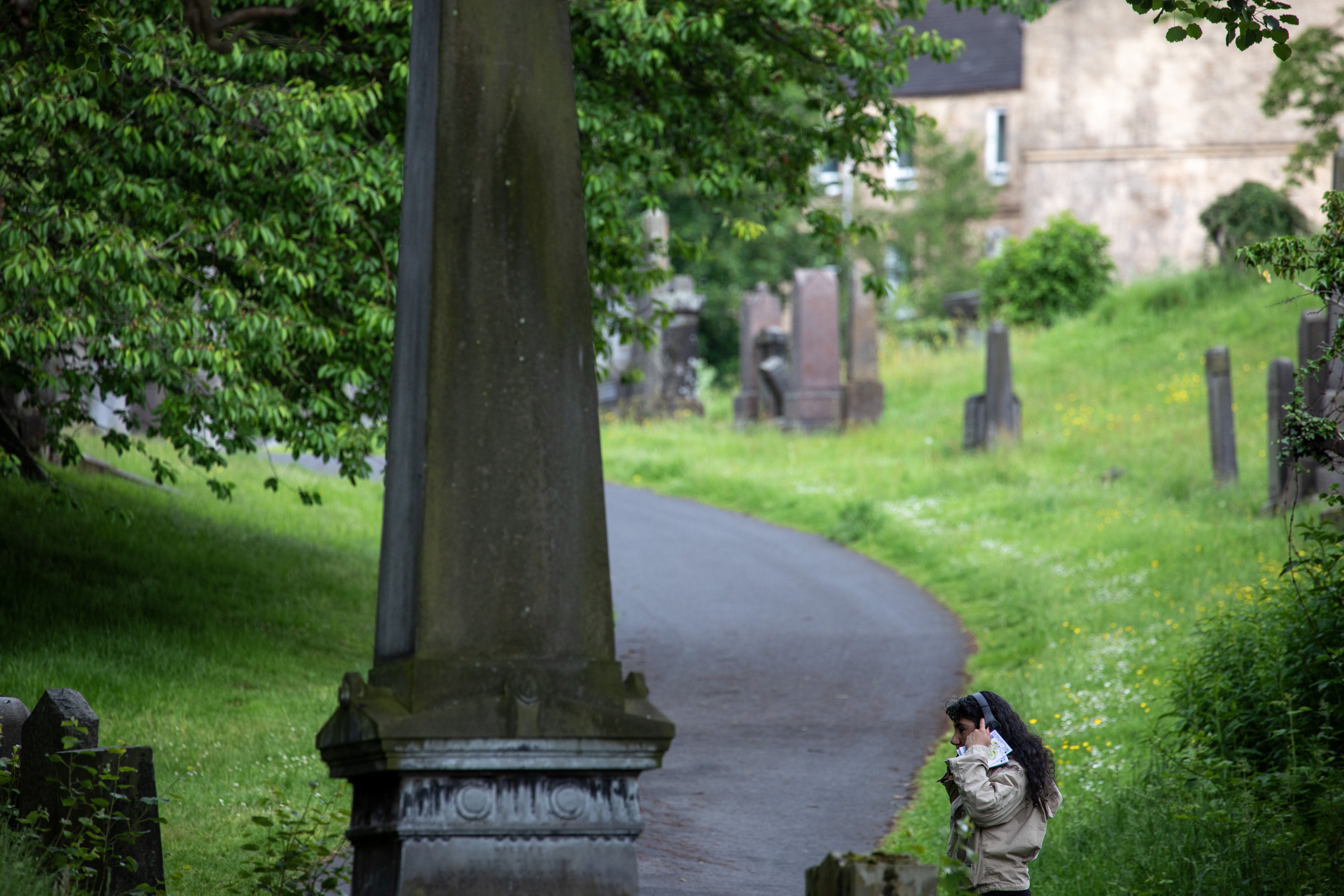 A person wearing headphones walks along a path through Glasgow Necropolis, following a printed map, with a large obelisk in the foreground and historic tenement buildings visible beyond the graves.