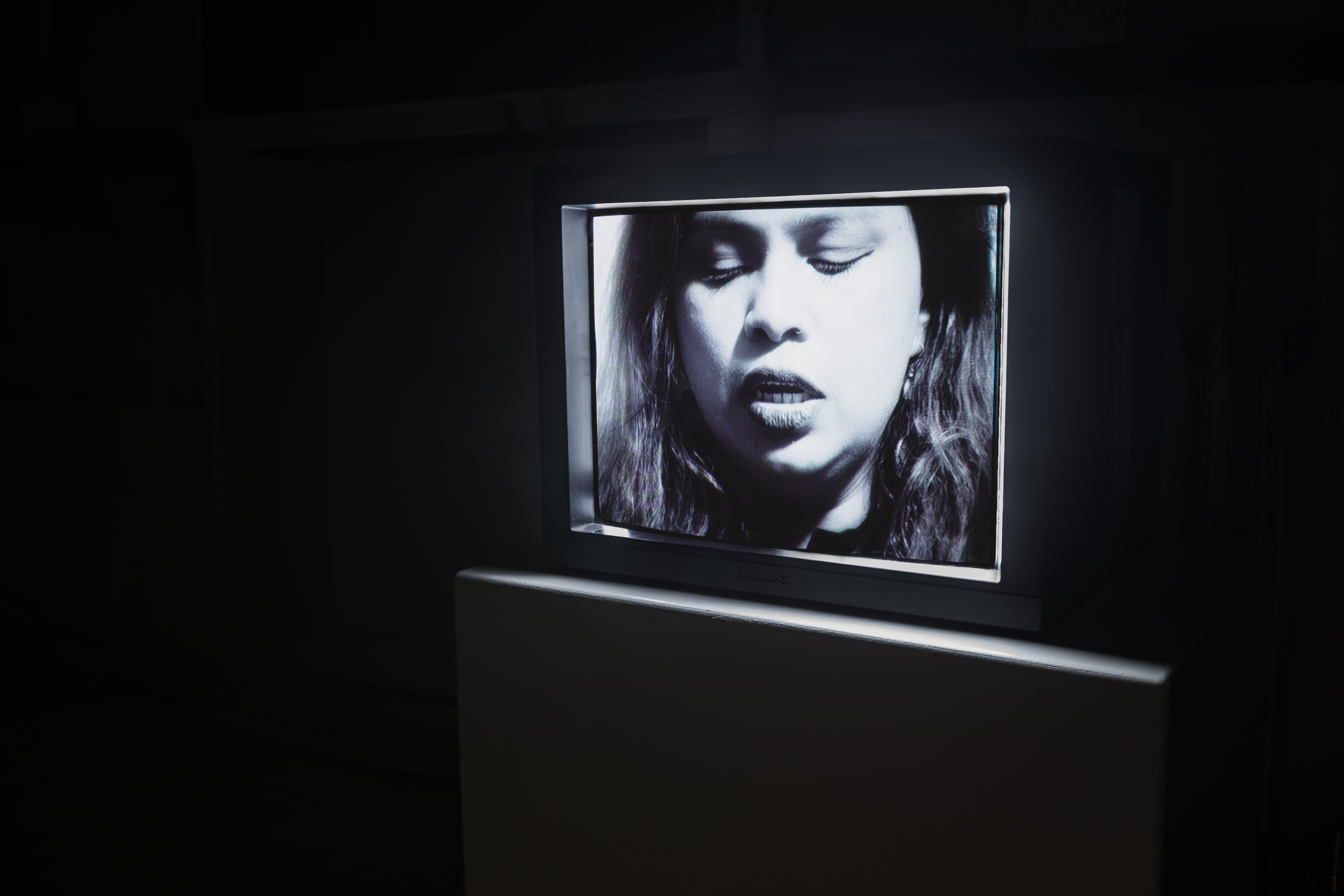 A vintage Sony CRT television glowing in a dark gallery space, displaying a black-and-white close-up of a woman singing with her eyes closed.