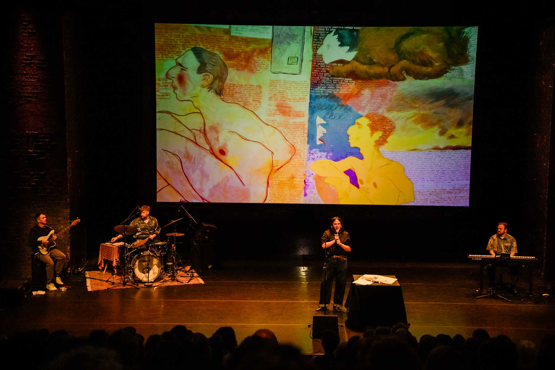 A performer stands at a microphone with musicians on stage beneath a large projected artwork featuring colorful illustrated figures and text.