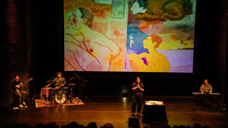 A performer stands at a microphone with musicians on stage beneath a large projected artwork featuring colorful illustrated figures and text.