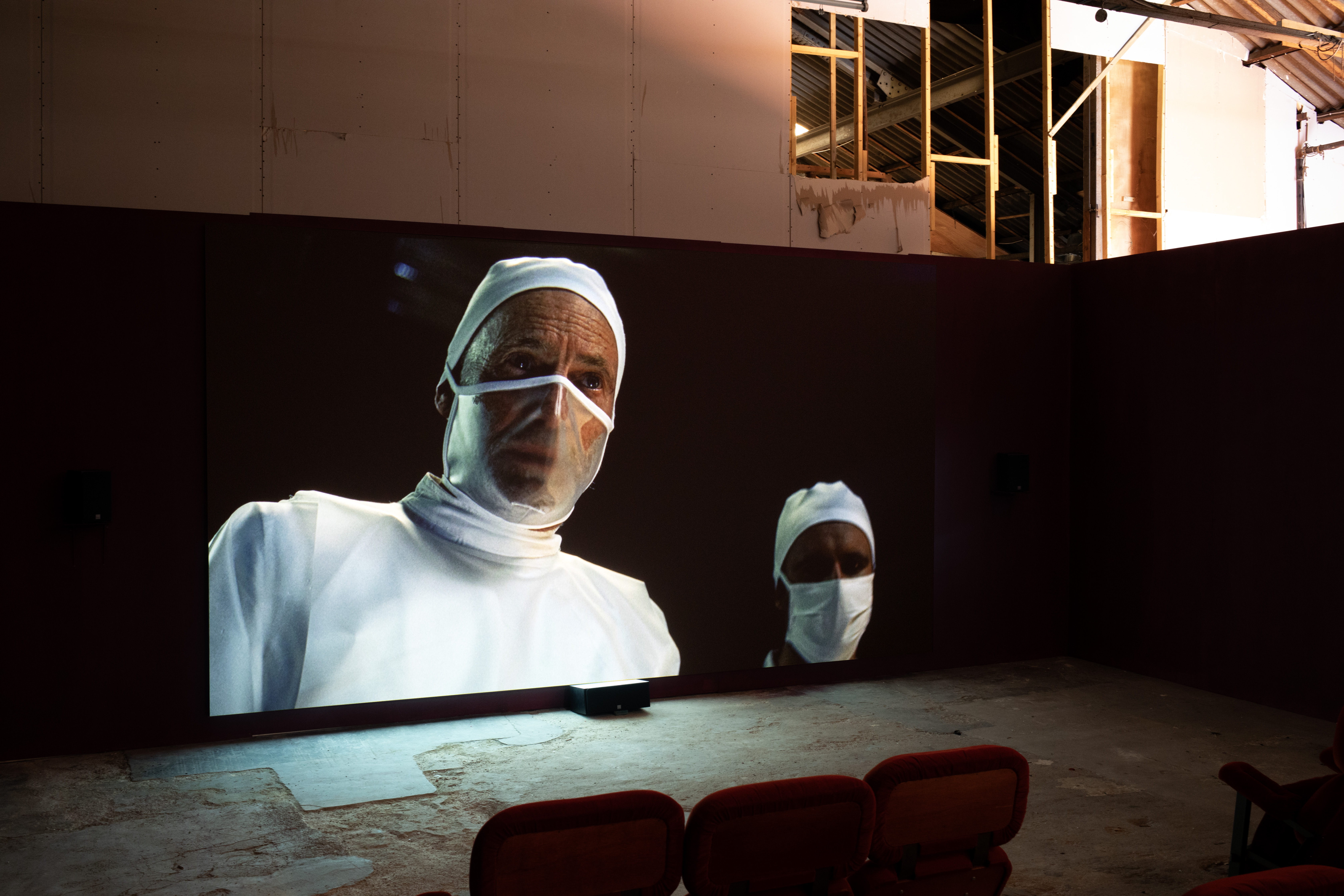A wide screen in a warehouse gallery projecting a still from Michelle Williams Gamaker's film "Strange Evidence," showing two figures in surgical caps and masks, with red cinema-style seating in the foreground and exposed roof timbers above.
