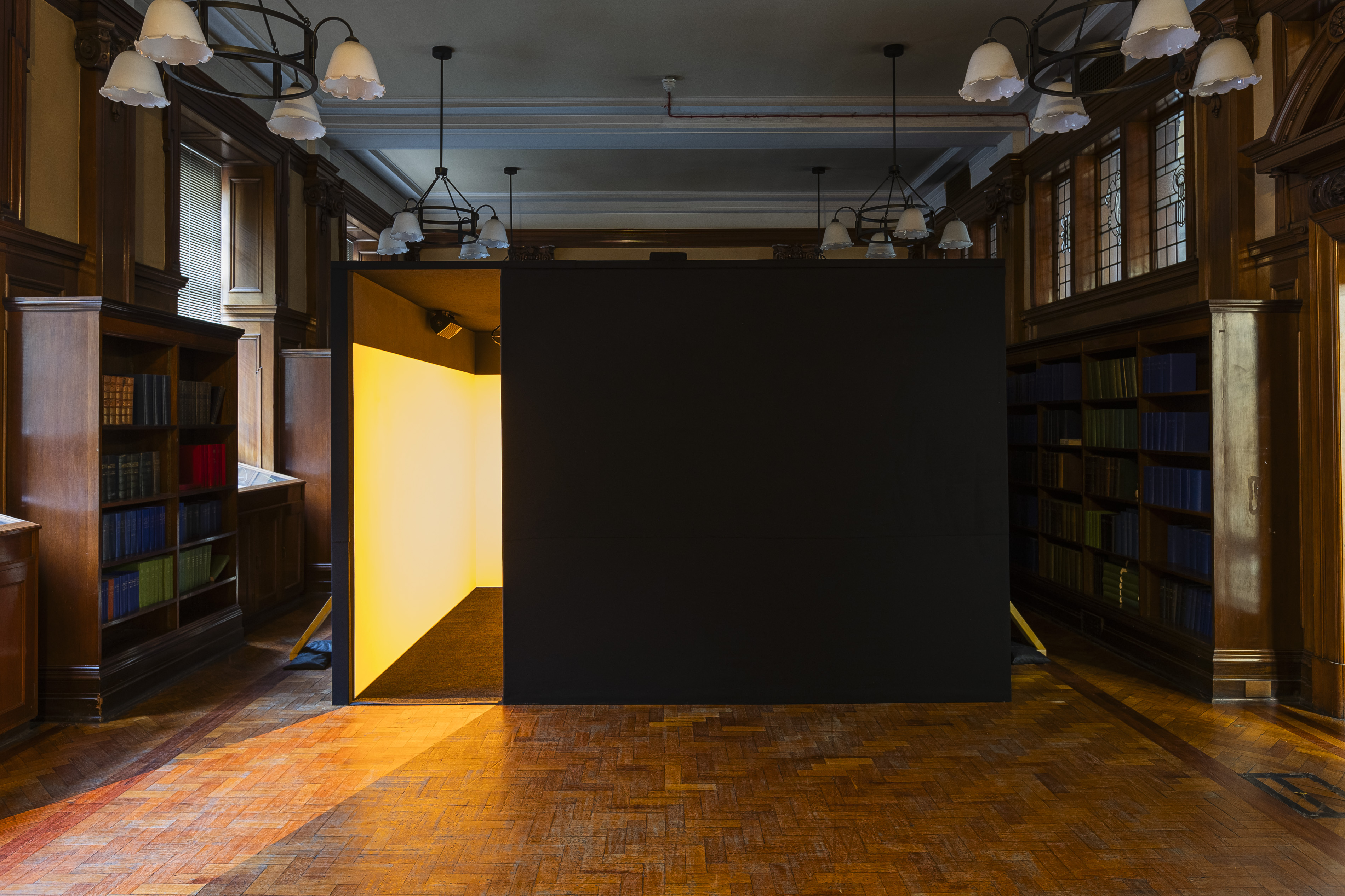 A dark walled structure with a glowing yellow interior stands in the Mitchell Library's wood-panelled reading room, part of Irienu Destourelles' installation, lit warmly against the surrounding bookshelves and pendant lamps.