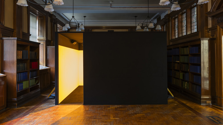 A dark walled structure with a glowing yellow interior stands in the Mitchell Library's wood-panelled reading room, part of Irienu Destourelles' installation, lit warmly against the surrounding bookshelves and pendant lamps.