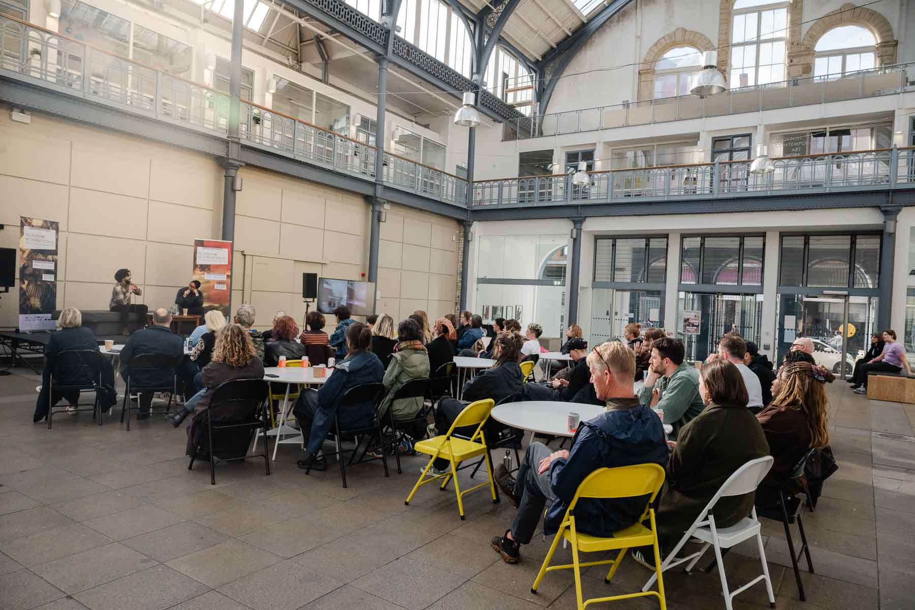 Wide view of an audience seated at round tables in a large glass-roofed hall, watching two people in conversation on a low stage with a screen displaying an image beside them.