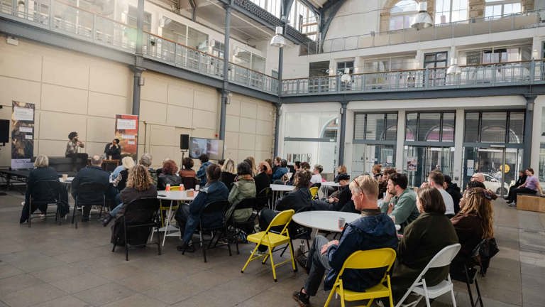 Wide view of an audience seated at round tables in a large glass-roofed hall, watching two people in conversation on a low stage with a screen displaying an image beside them.
