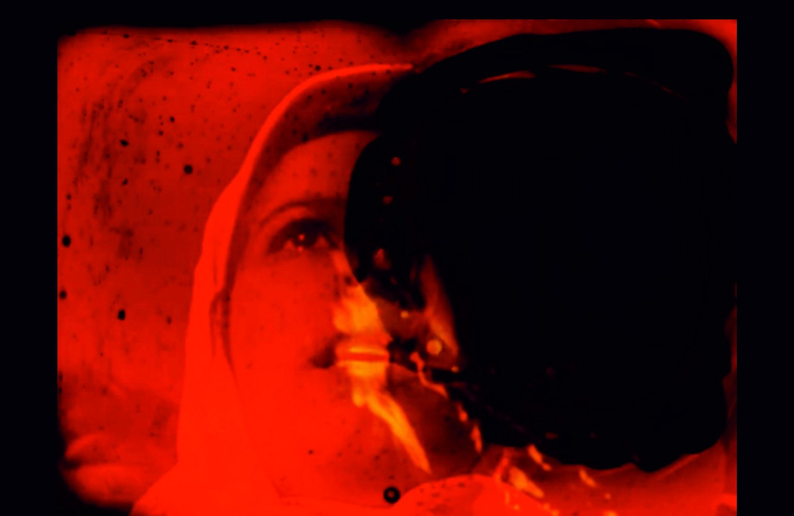A film still of a women wearing a bonnet. Her face shows fear or despair and is partially obscured by a dark mass, with flames visible in the background.