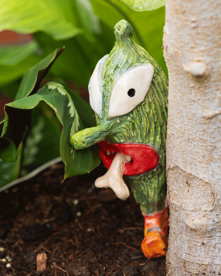 Close up view of a small ceramic sculpture in a plant pot of a green figure with red lips