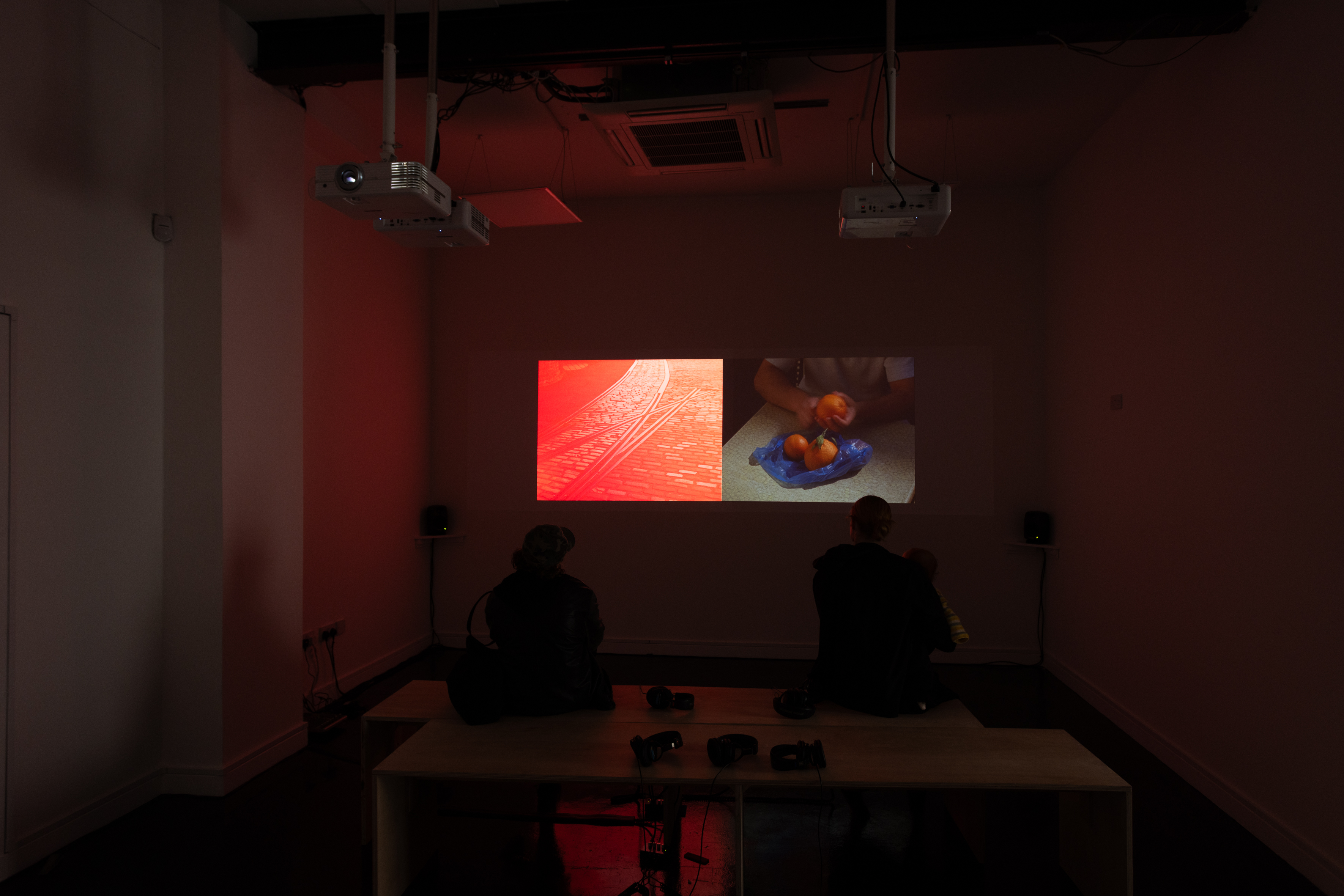 Wide installation view of a darkened gallery space with two ceiling-mounted projectors casting a split-screen video work onto the wall, showing red-toned cobbled tram tracks alongside footage of hands peeling oranges, with two visitors seated on a low platform watching, headphones laid out in front of them.