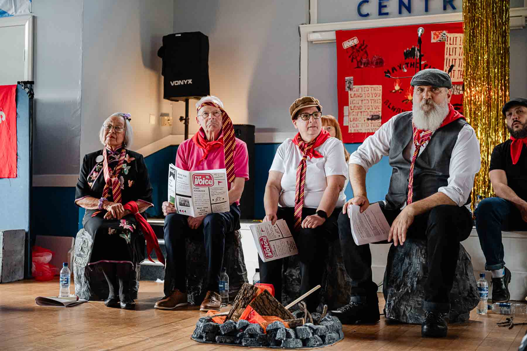 Four seated performers in red neckerchiefs look on attentively beside a prop campfire, with a "May the Fire Always Be Lit" banner and gold tinsel curtain behind them.