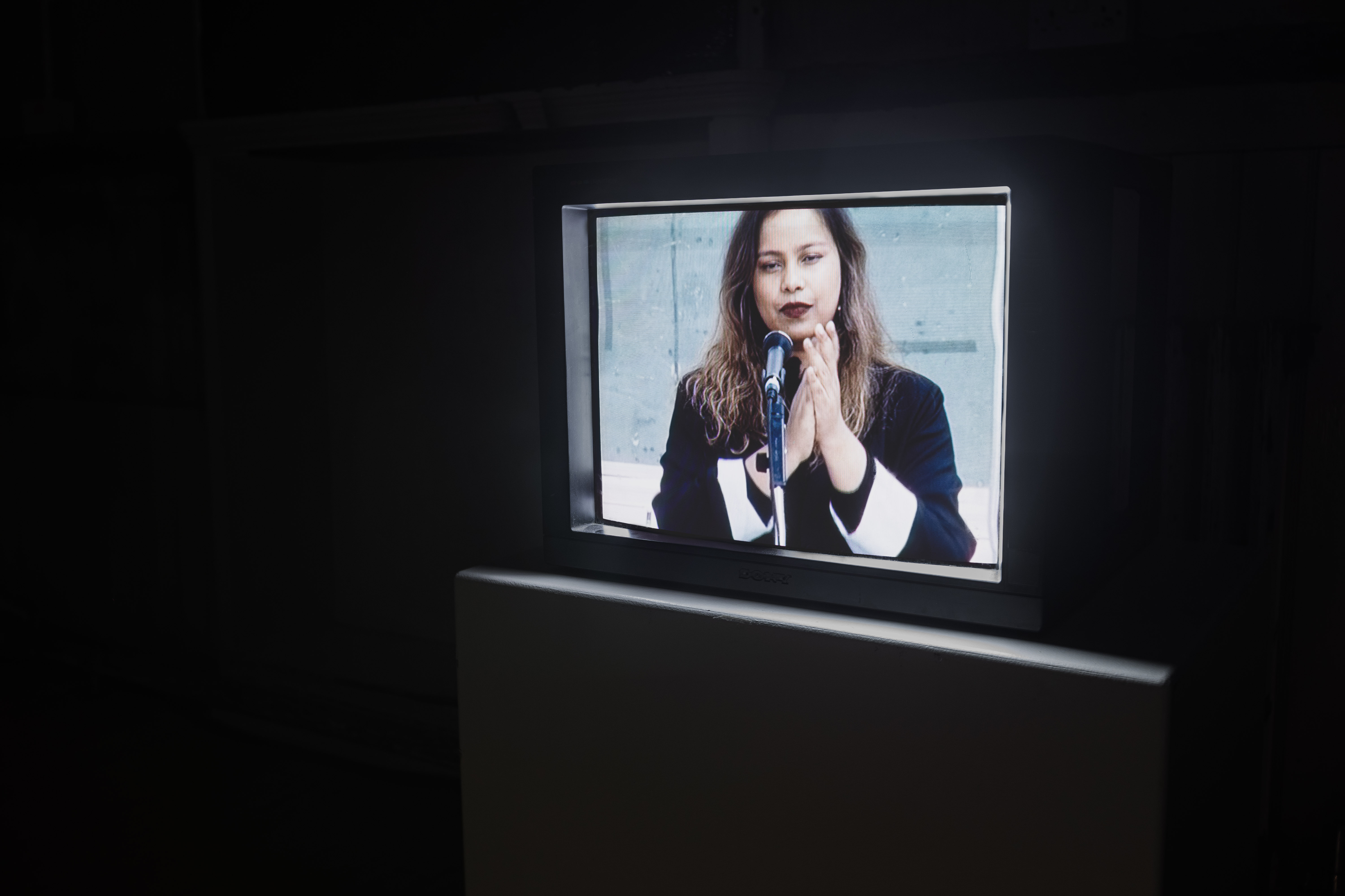 A vintage Sony CRT television in a dark gallery displaying a colour image of a woman with clasped hands standing at a microphone, wearing a black and white jacket.