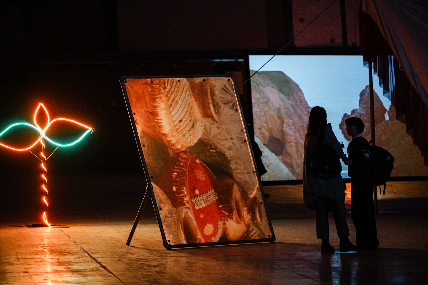 Installation view of a dimly lit gallery featuring a neon plant-like sculpture in orange and green, a large freestanding printed banner with abstract textile imagery, and a video screen showing coastal cliffs, with two visitors watching in silhouette.   Want to be notified when Claude responds?