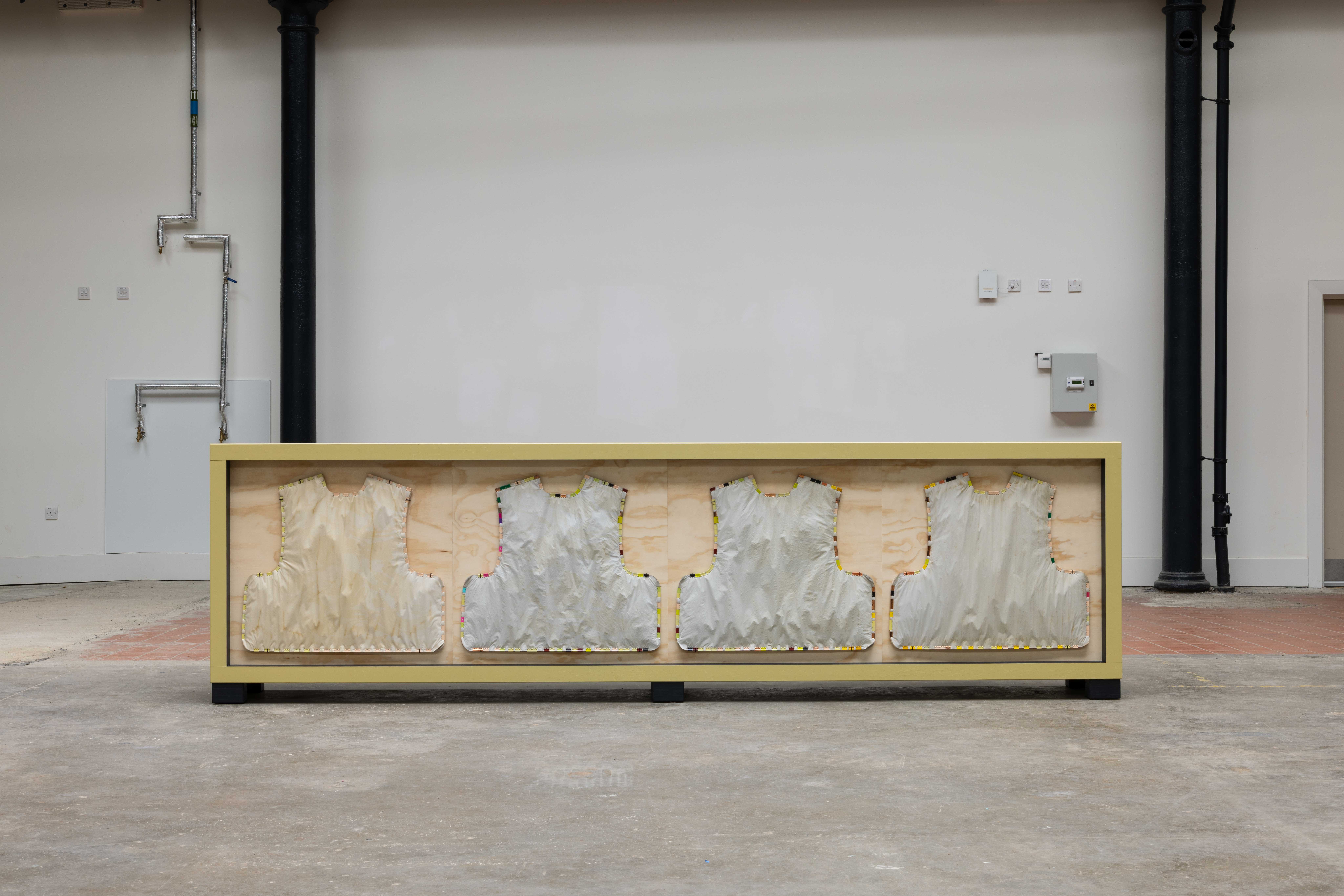 Long yellow-framed wooden crate sculpture displaying four vest-shaped fabric forms mounted side by side, set against a plain gallery wall with exposed pipework and a fire escape door visible.