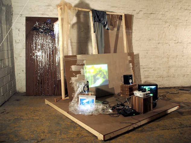canvas frames, plywood, and scrap material leaned against each other. beneath in the floor is a crt monitor, two small plinths, a projector, speaker, and some other technical equipment.
