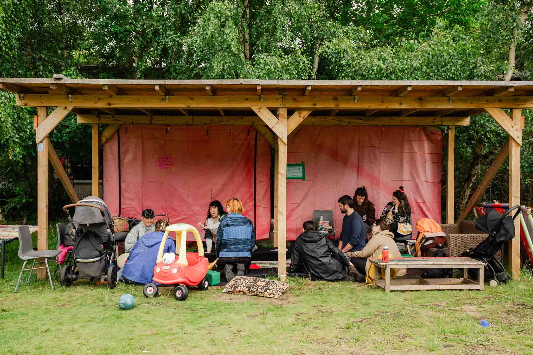 A group of adults and children gather under a wooden shelter with pink fabric walls, sitting on benches and cushions surrounded by pushchairs and a toy car on the grass.