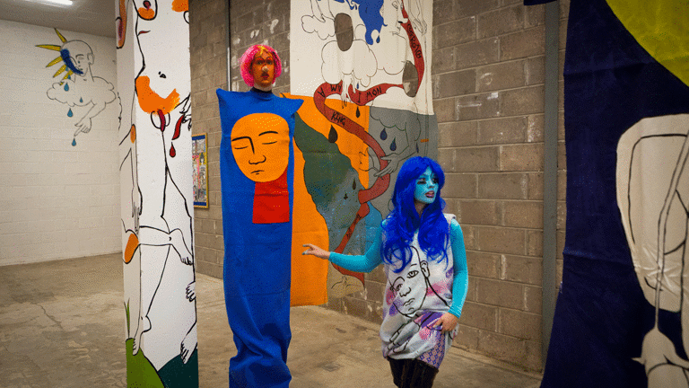 Playful and surreal line drawings of figures with blocks of colour cover a wall, column and fabric hanging in an industrial space. There are two performers, one appears to be extra tall and is wearing a tabard-like column of blue fabric with a disappointed looking cut out of an orange face, the performer's face is painted orange and they wear a red wig. The other performer has a turquoise painted face and electric blue wig, the line drawings of the wall cover their clothing.