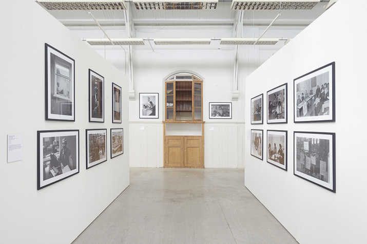 Photographs are displayed on two parallel walls of a white room. In the centre is a wooden cabinet.