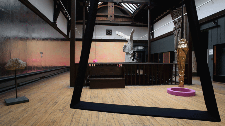 Behind a large suspended rectangular frame, there is a metallic vertical sculpture on a pink ring shaped stand. There is another sculpture on a stand on the left side and another at the back of them room. The one at the back of the hallway looks like an angel. 