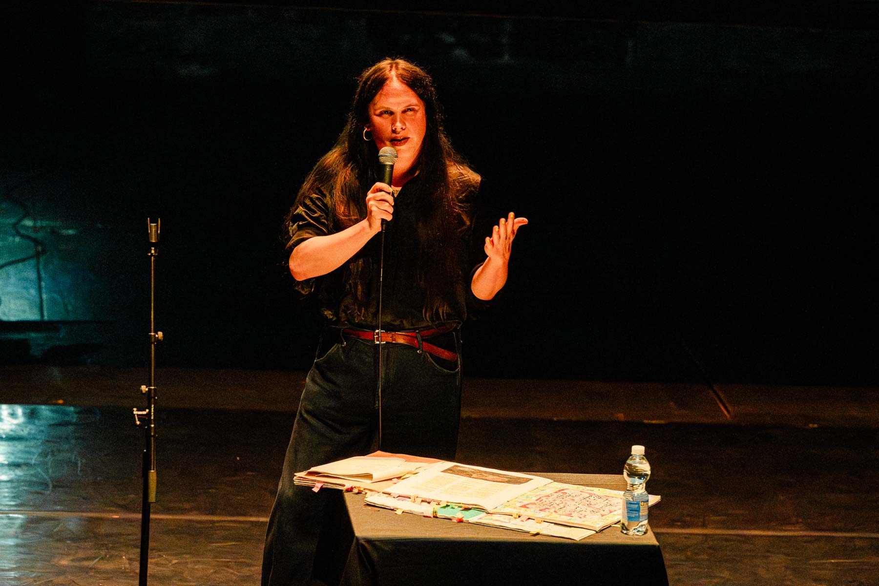 A person speaks into a handheld microphone on a dimly lit stage beside a table covered with open books, papers, and a water bottle during a live presentation.