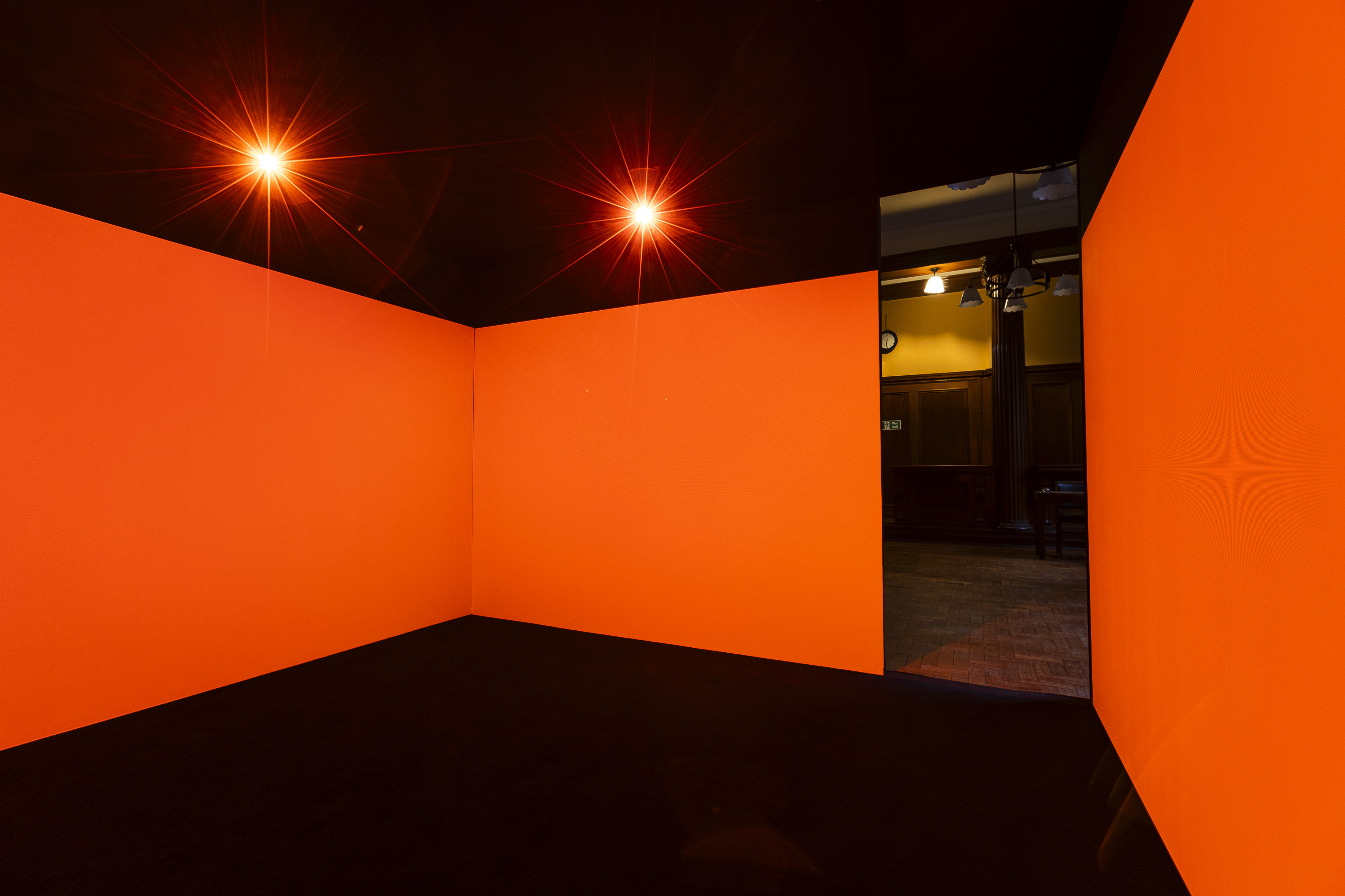 Inside Irienu Destourelles' installation, an empty room with vivid orange walls and black ceiling features two intense starburst light sources, with a glimpse of the Mitchell Library's panelled room visible through a doorway.
