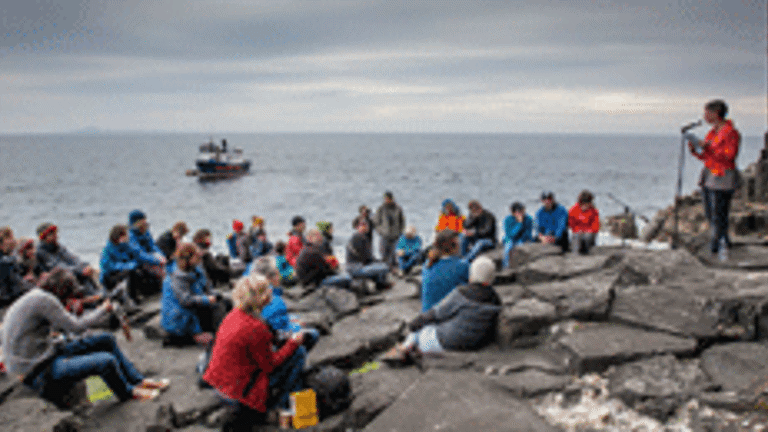 A photograph of a large group of people sitting on a rocky shore with sea in the back ground and boat. A person is reading to the group, speaking through a microphone