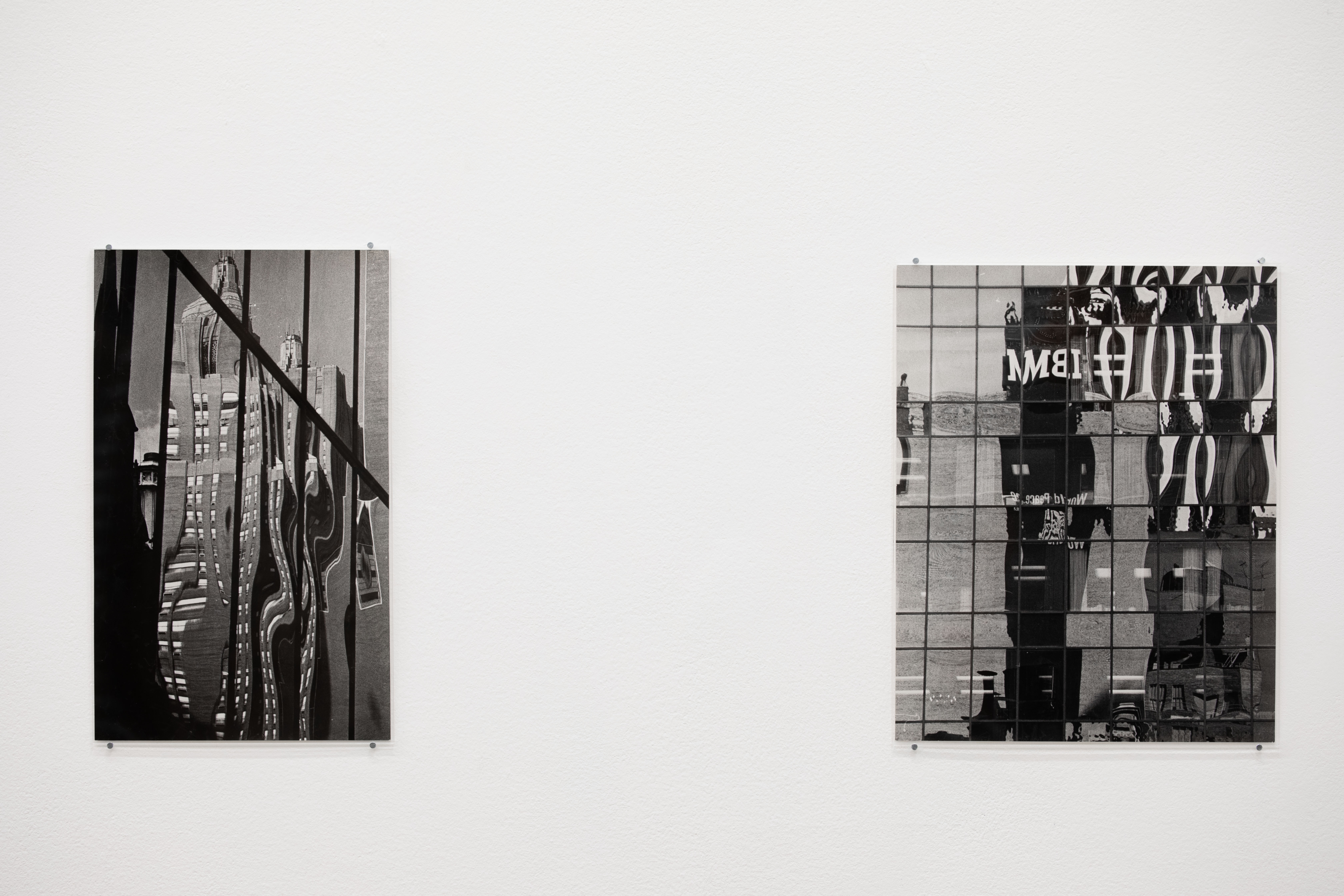 Two black-and-white photographs mounted on a white gallery wall, both showing distorted reflections in glass-panelled skyscraper facades, one bearing a mirrored "IBM" logo.